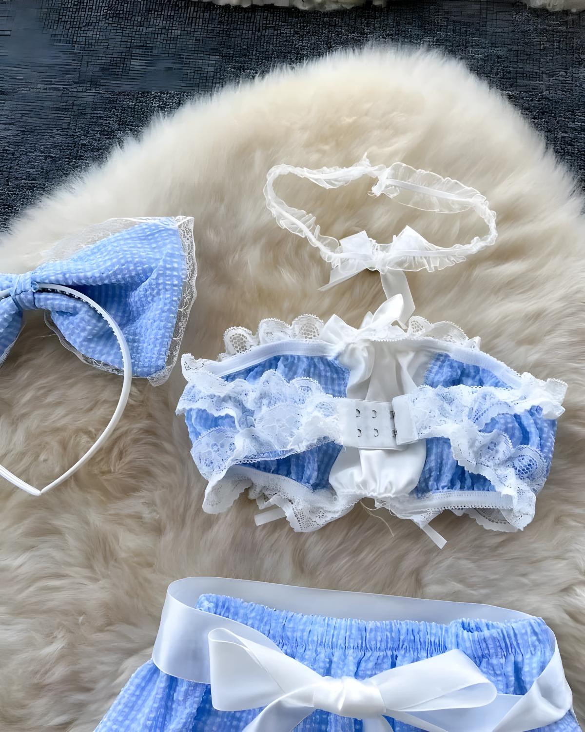A two-piece babydoll lingerie set in light blue and white, featuring a ruffled lace bralette with a white lace collar, matching blue and white lace garter belt, and a large white bow on the waistband, displayed on a fluffy cream faux fur surface.