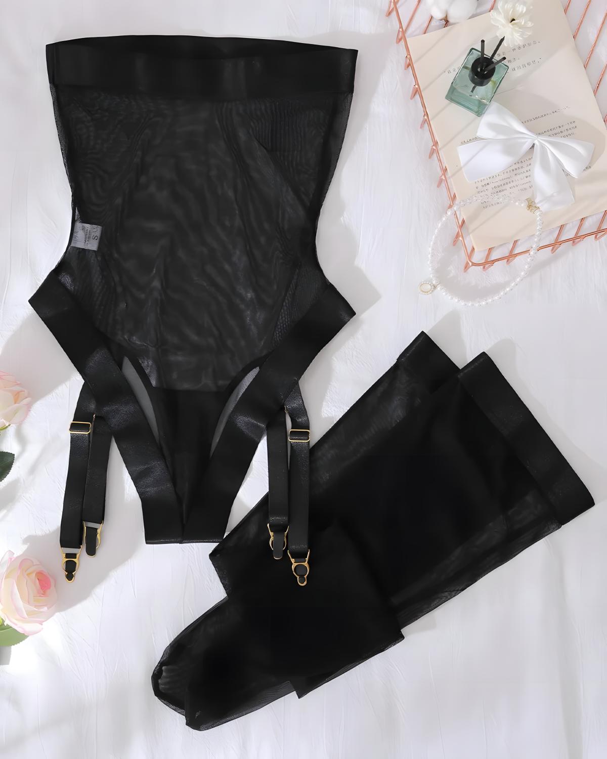 Black sheer mesh and satin suspender garter belt set with attached thigh straps and gold-tone hardware, arranged on a white fabric surface with decorative flowers and a small gift box.