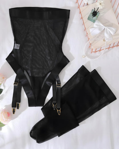 Black sheer mesh and satin suspender garter belt set with attached thigh straps and gold-tone hardware, arranged on a white fabric surface with decorative flowers and a small gift box.