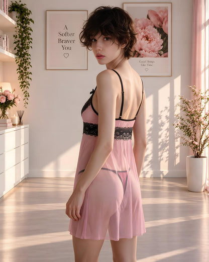 A woman stands in a sunlit bedroom wearing a light pink babydoll chemise with black lace trim and black spaghetti straps, photographed from the back.