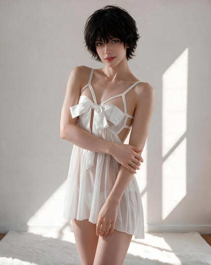 A woman modeling a white sheer babydoll lingerie dress with a large bow at the bust, photographed in a bright room with sunlight and shadows.