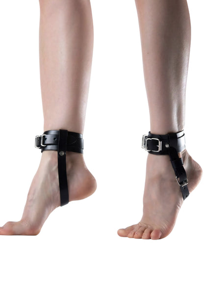 Black ankle cuffs with silver buckles worn on both ankles, shown on a person's feet against a plain white background.