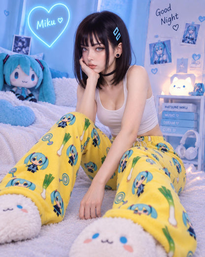 A woman sits on a bed in a pastel bedroom setting, wearing bright yellow plush pajama pants covered in cute blue anime character prints, a white tank top, and fuzzy white slippers. The room has blue lighting, a plush toy, and decorative posters.
