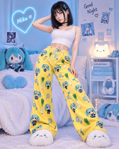 A model stands in a bedroom setting wearing bright yellow wide-leg pajama pants covered in a repeating character and green vegetable print, paired with a white crop top and fuzzy white slippers. The room is decorated with blue-themed anime props, including a plush character, a glowing heart sign that says 'Miku', and shelves with books and headphones.