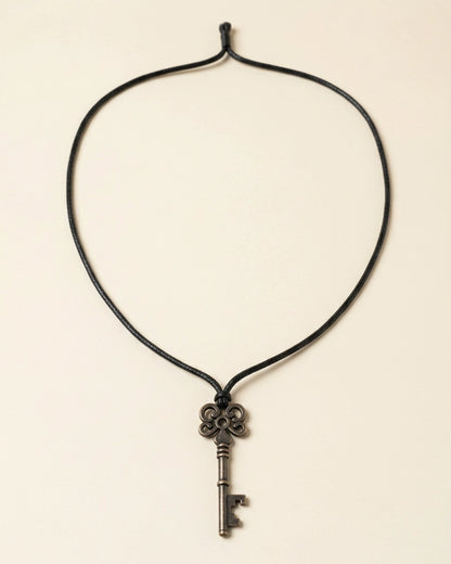 A black cord necklace featuring an antique-style metal key pendant, displayed against a solid light beige background.